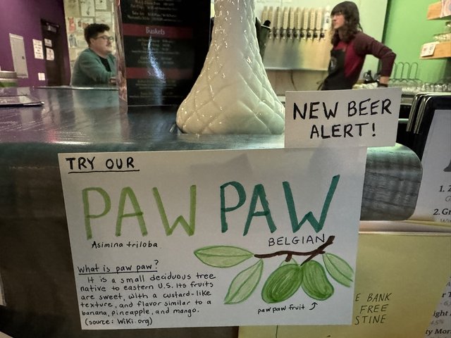 Handwritten sign advertising Paw Paw beer, with taproom and two person s blurred in the background.