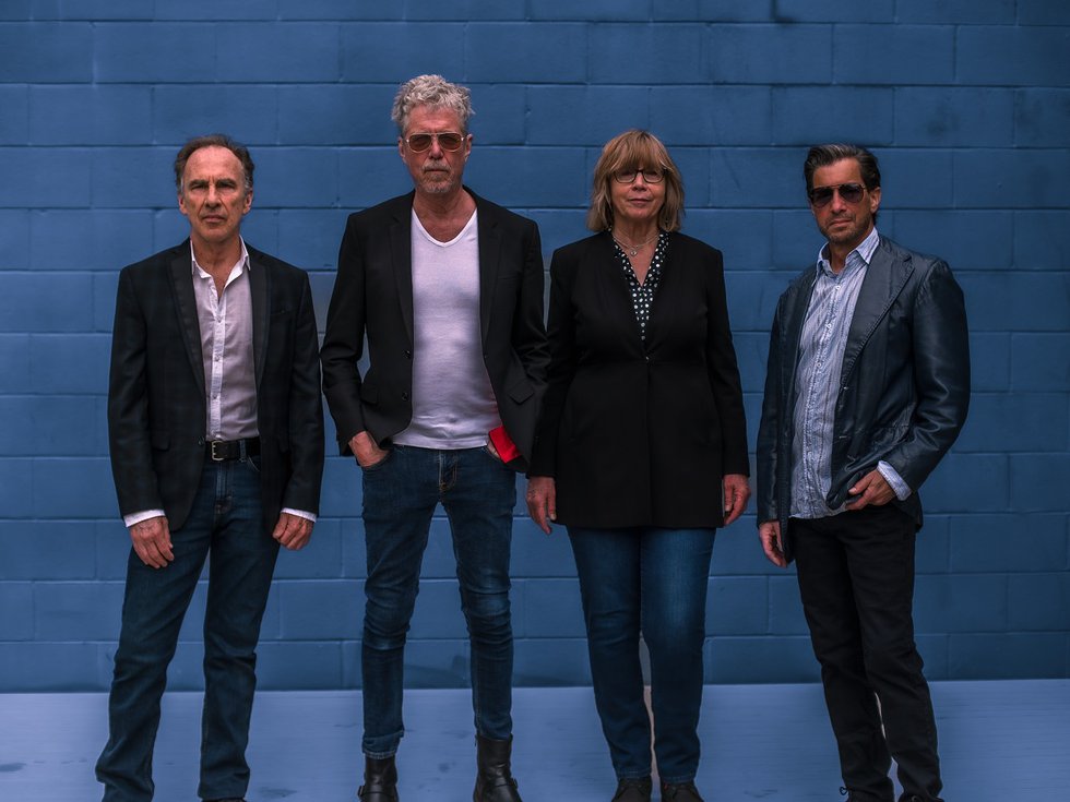 The four members of The Jayhawks in front of a blue wall.