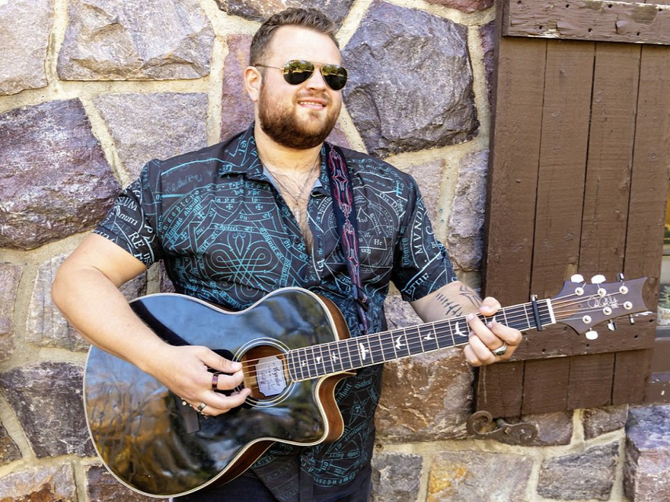 Taylor Schereck and guitar in front of a stone wall.
