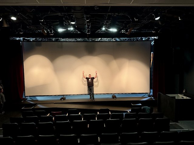 A man standing in front of a large movie screen holding his hands up in the air.
